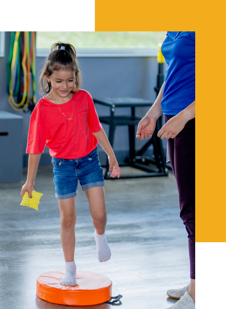 Child practicing balance on a therapy cushion with guidance from a physical therapist in a clinic setting.