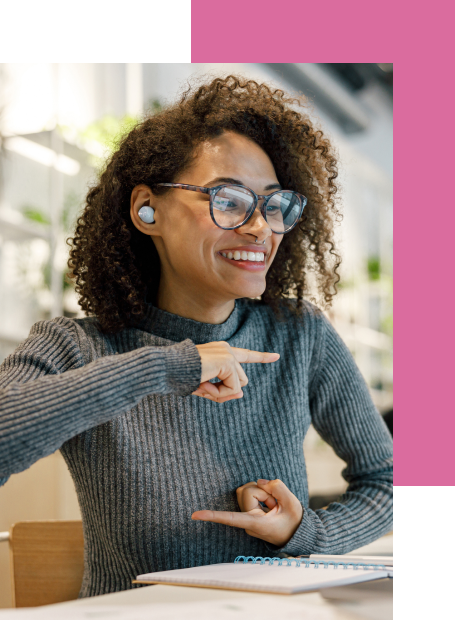 Remote sign language interpreter communicating via video, gesturing while seated at a desk with a notebook.